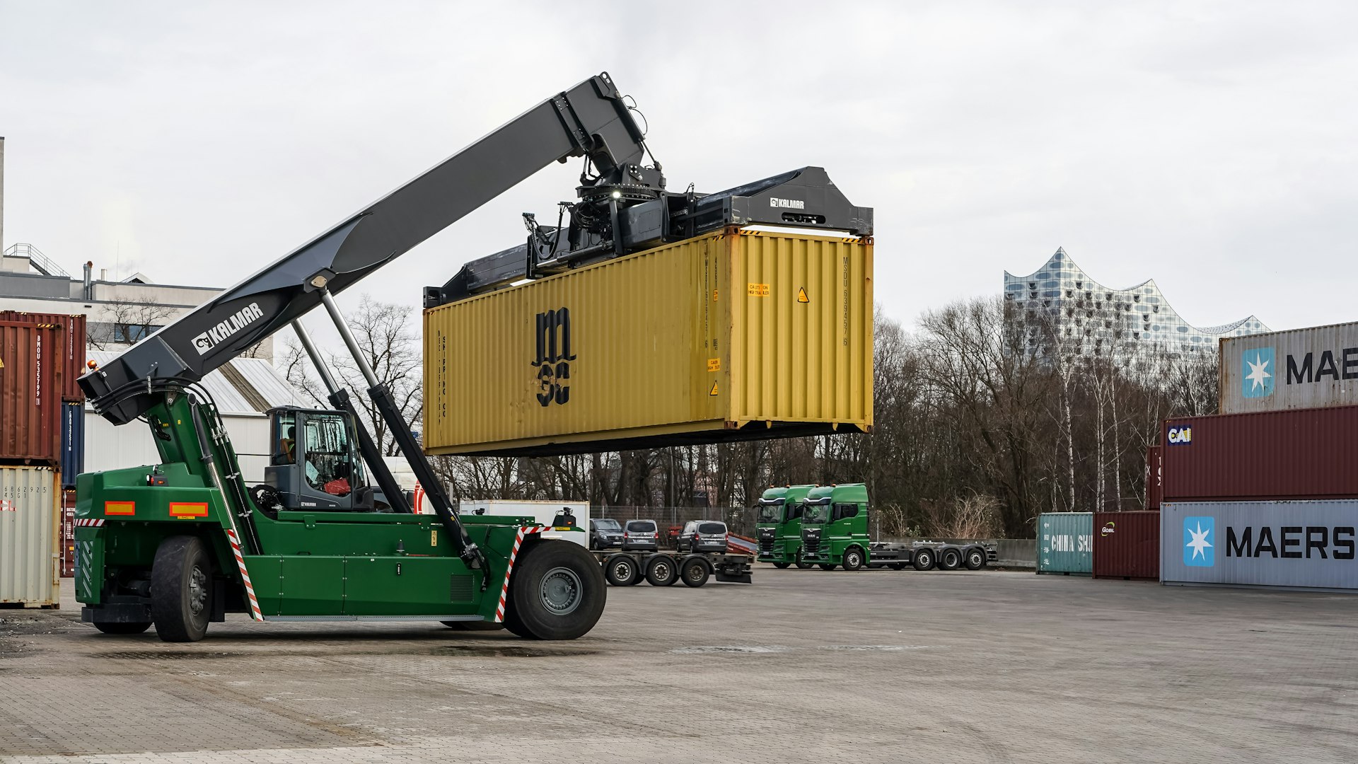 A green reach stacker lifting a yellow shipping container.